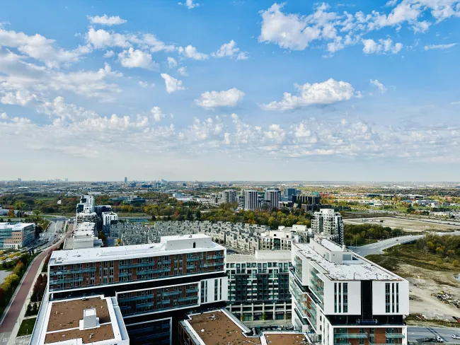 A high-angle aerial view of a modern urban neighborhood with mid-rise apartment buildings and roads in the foreground, autumn-colored trees and a sprawling cityscape beyond, under a bright blue sky filled with scattered white clouds.