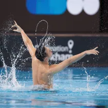 A synchronized swimmer performs a dramatic routine with arms extended in a pool, creating water splashes.