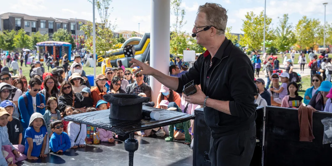 A performer on an outdoor stage demonstrates a magic trick beside a top hat while a large crowd of adults and children watches from the audience.
