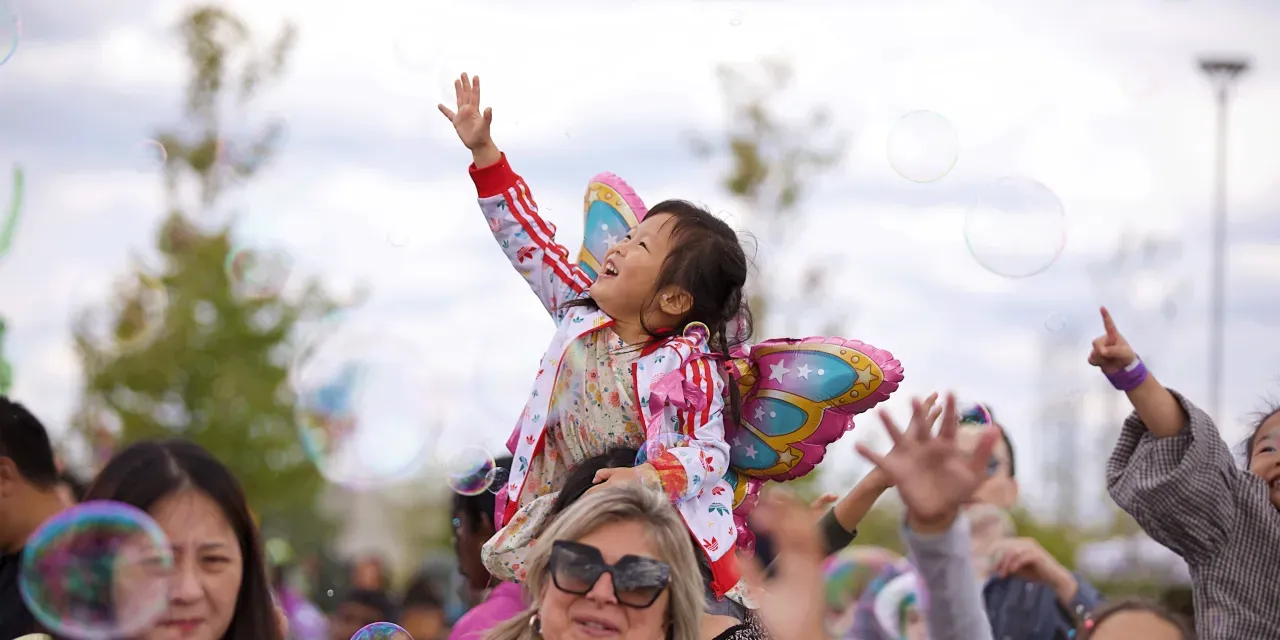 Child with butterfly wings sitting on parent's shoulders reaches for bubbles in the air