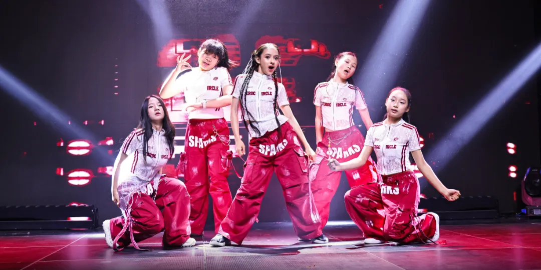 Five Soul Attention Dance School performers in matching white tops and red pants pose in a dance formation on a stage, illuminated by bright spotlights and a red-lit backdrop.
